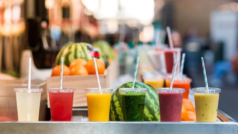 Six clear plastic cups full of colourful juices sit in front of various fruits.