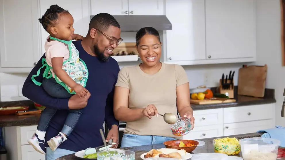 A family of three by the kitchen table, with the mother preparing the meal and the father holding their child in one arm.