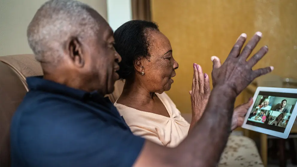 An older black couple sit on the couch and speak to their friends through a video call on a handheld tablet. 