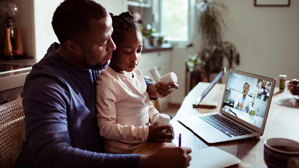 A father with his daughter on his lap attending a virtual doctor’s appointment. 