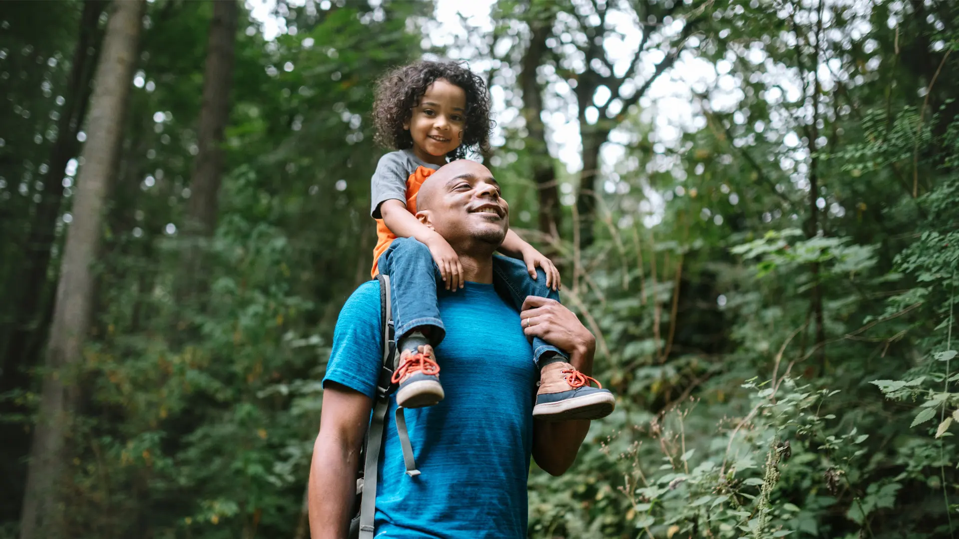 A man walks through a trail while his young daughter sits on his shoulders