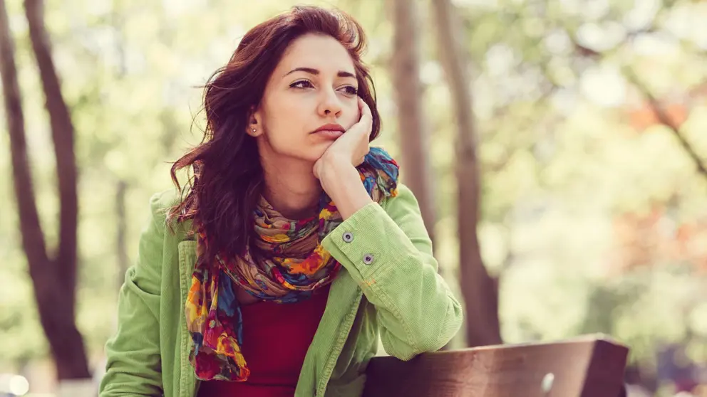 A woman sits outdoors with a hand on her chin.