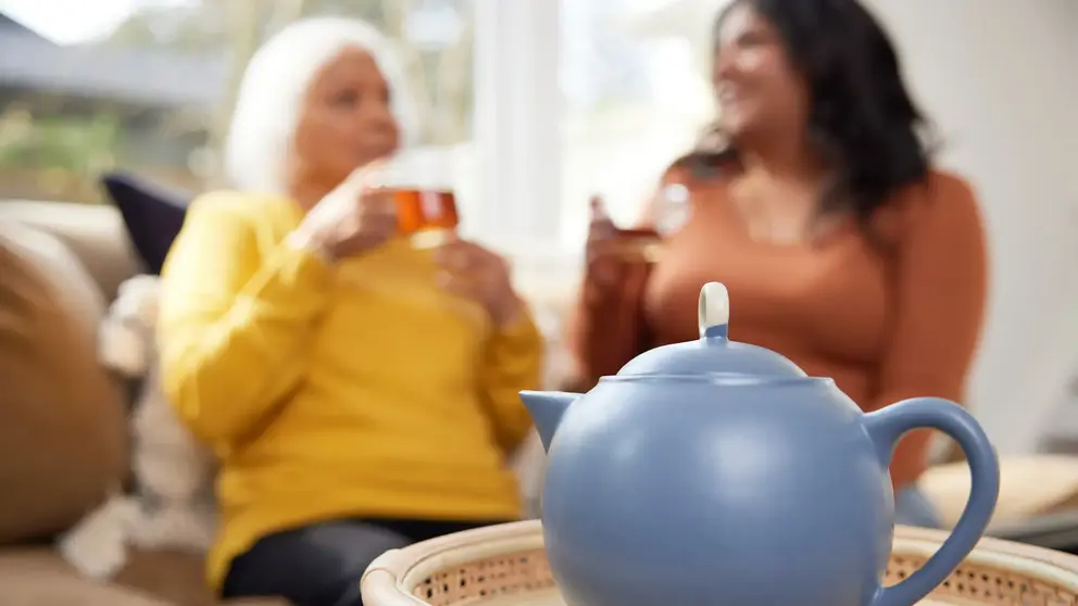 A blue teapot with two women drinking tea in the background.