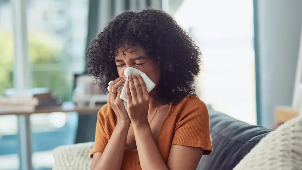 A woman blows her nose into a napkin while seated on a couch.