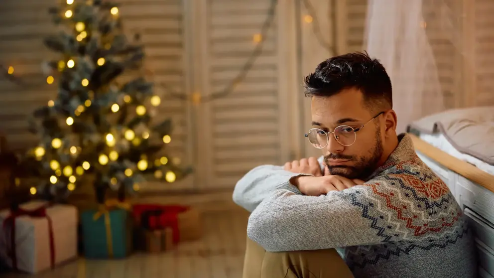 A man with a sad expression sits by the bed with a Christmas tree and presents in the background