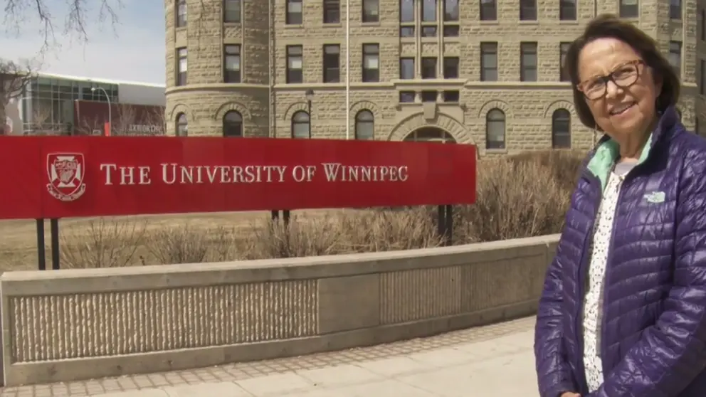 Esther Sanderson wears a purple jacket and smiles beside a University of Winnipeg sign.