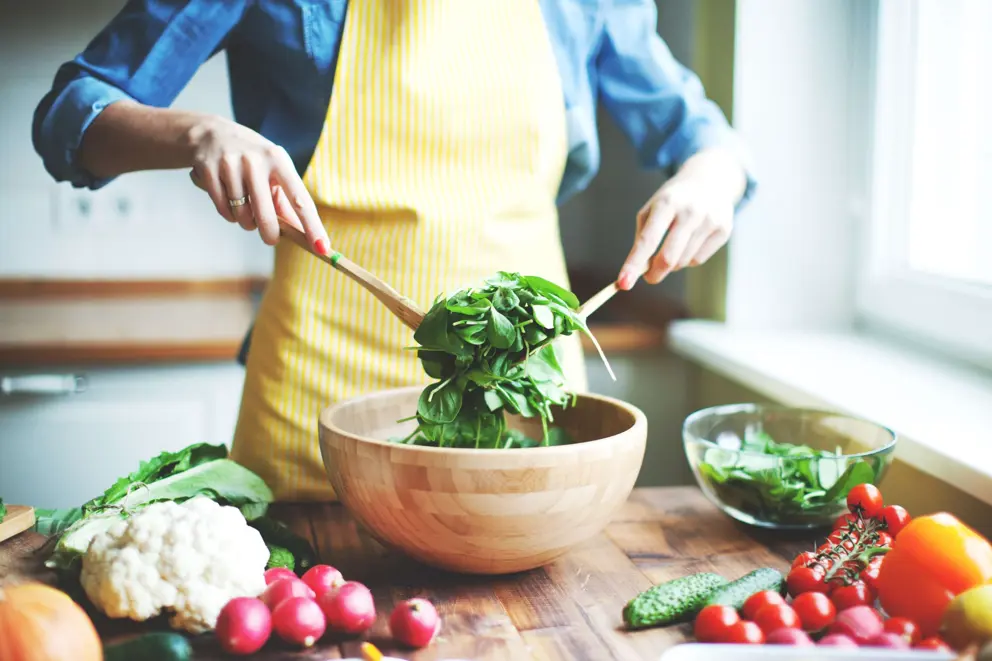 A woman wearing a yellow apron tosses green salad in a wooden bowl.