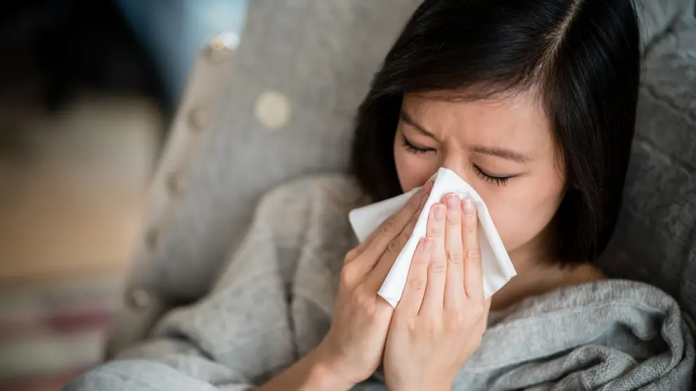  A woman wearing a gray sweater blows her nose into a tissue.