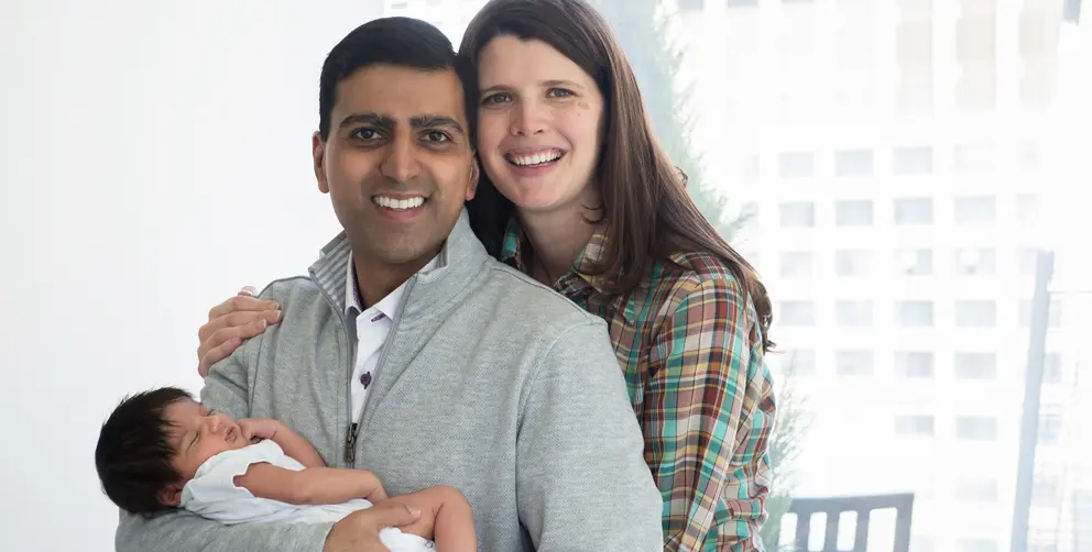 Irfhan Rawji and his wife Christine, stand close together with arms wrapped around their new born baby boy, Zain, while smiling at the camera. 