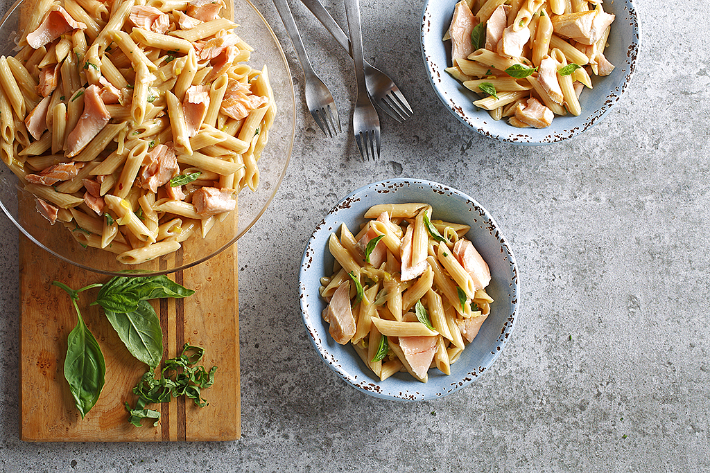 A glass serving dish of Salmon and leek pasta, beside two portions in gray ceramic bowls.