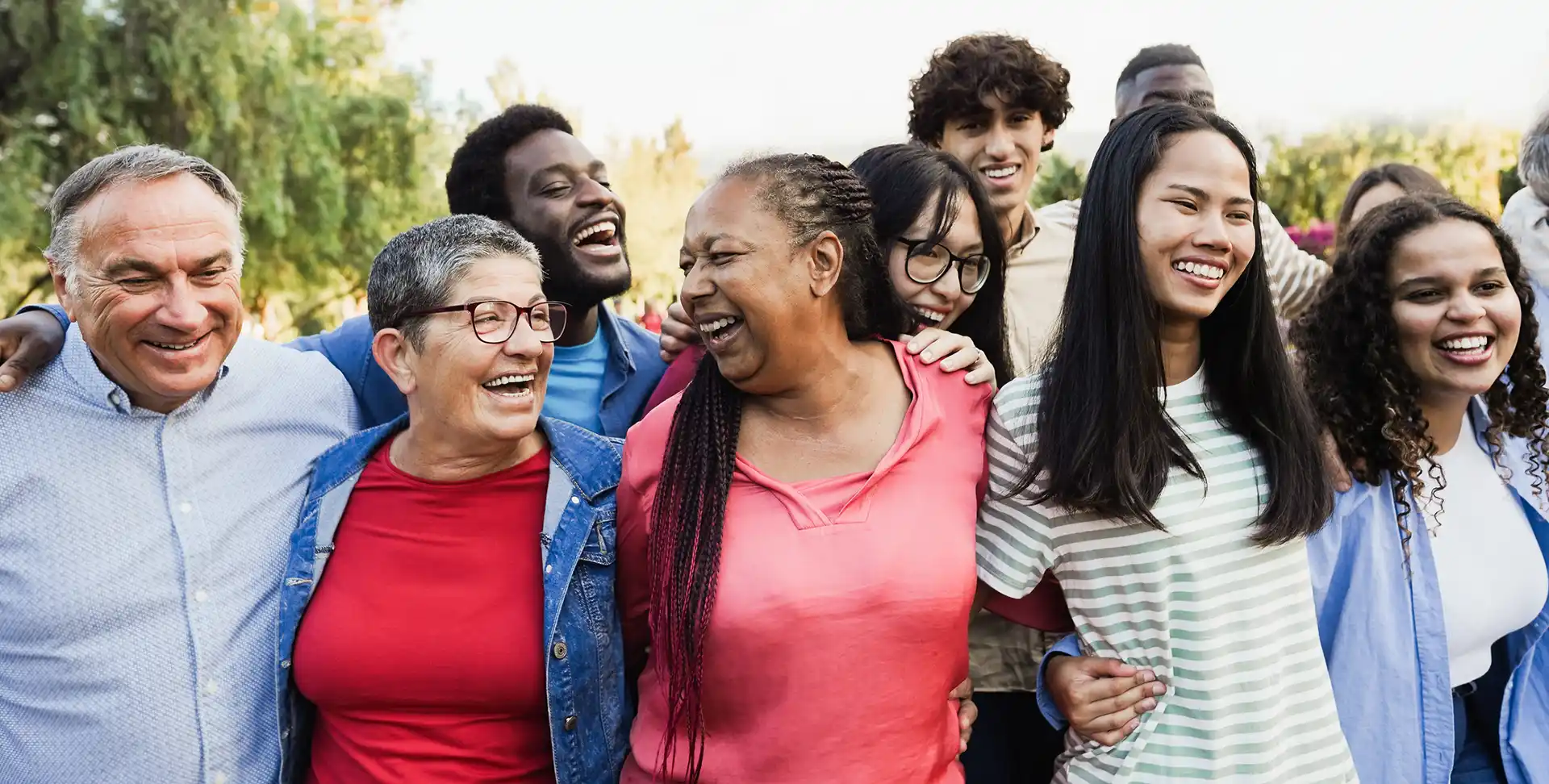 A large group of diverse people stand outdoors while smiling and holding each other.