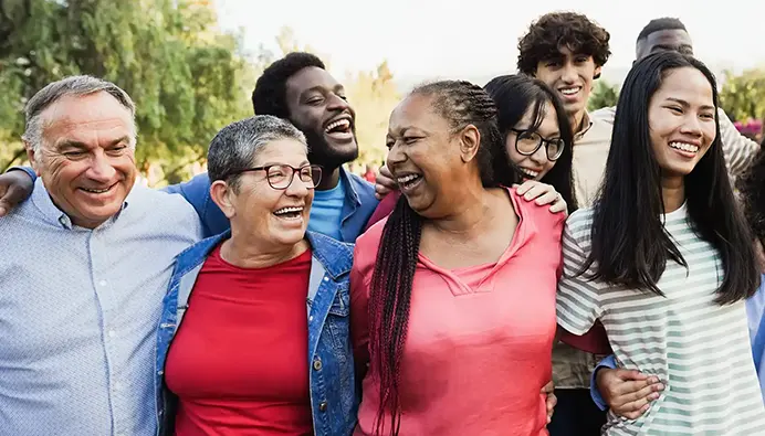 A large group of diverse people stand outdoors while smiling and holding each other.