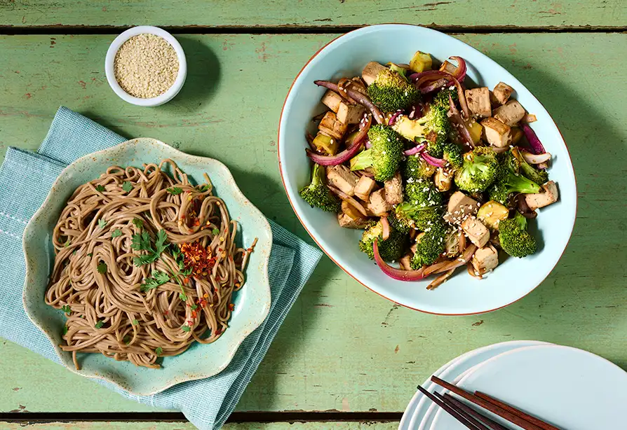 Tofu, broccoli and onion in a serving dish beside a bowl of noodles